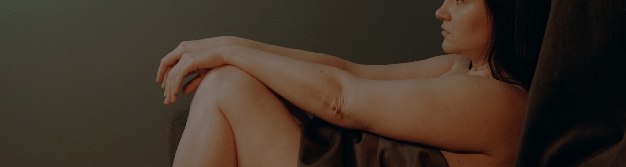 Eine Person sitzt seitlich auf einem Sofa, die Knie angezogen und die Arme darum gelegt. Die Beleuchtung ist gedämpft, was eine ruhige Atmosphäre schafft.