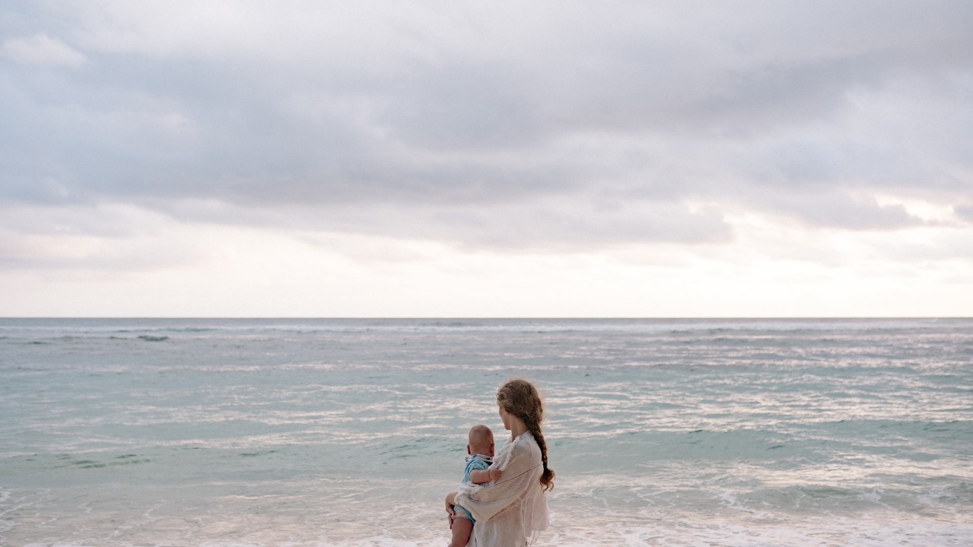 Eine Frau mit langen Haaren steht am Strand und hält ein kleines Kind im Arm. Beide blicken auf das ruhige Meer, während der Himmel bewölkt ist.