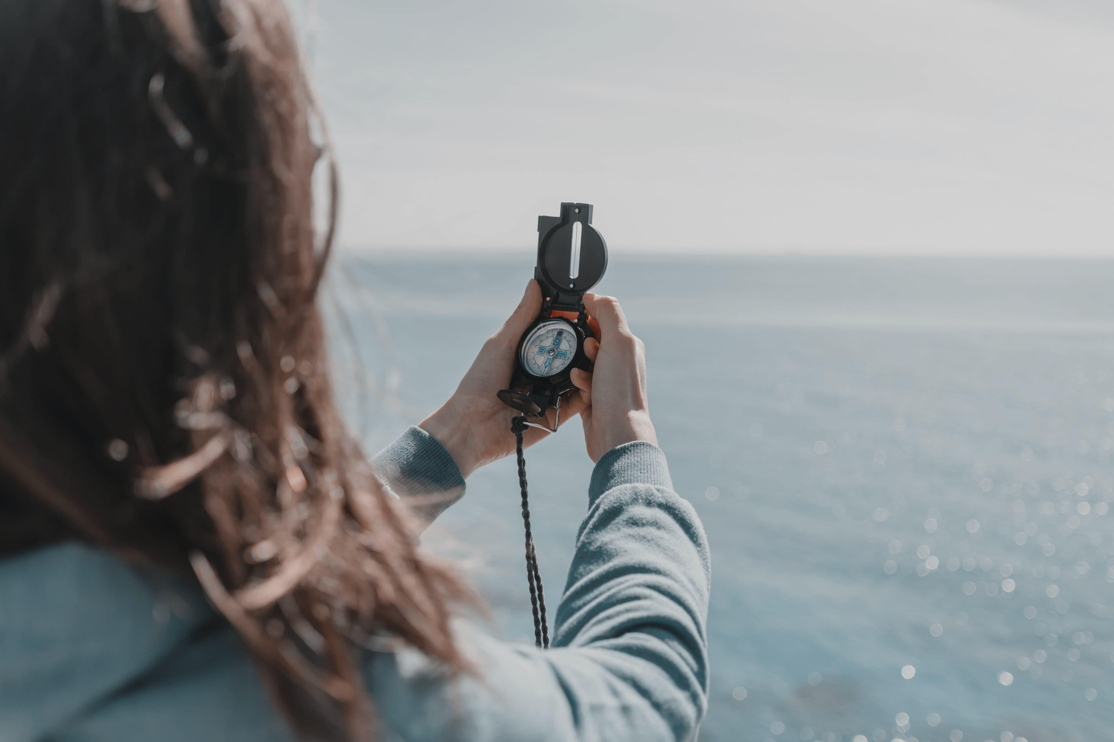 Eine Frau hält einen Kompass in der Hand und blickt auf das Meer. Die Szene ist von hinten aufgenommen, sodass die Person und der Kompass im Vordergrund sind, während das Meer im Hintergrund zu sehen ist.
