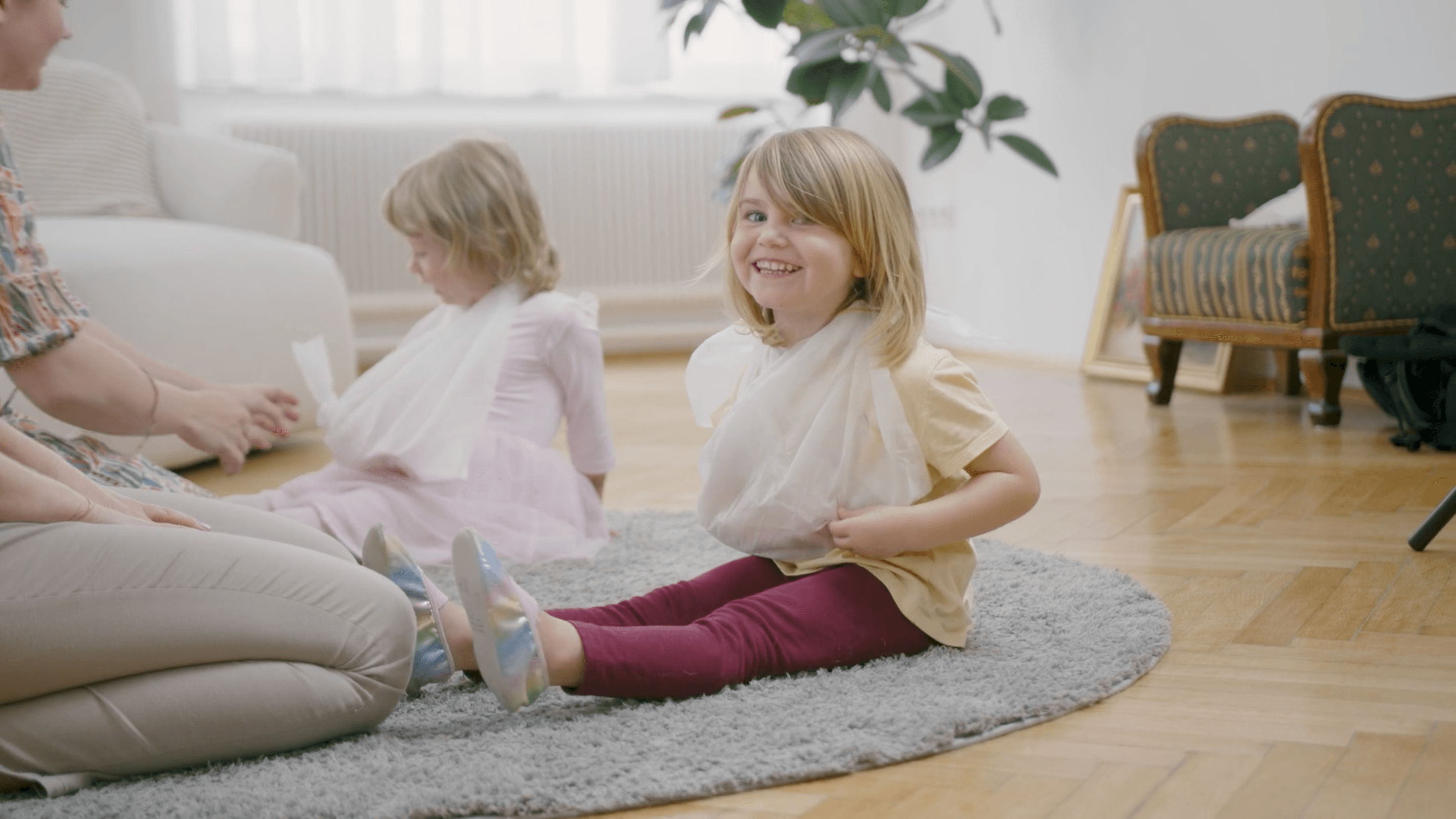 Zwei Kinder sitzen auf einem Teppich, eines lächelt fröhlich. Beide tragen Tücher um den Hals. Im Hintergrund sind Pflanzen und Möbel.